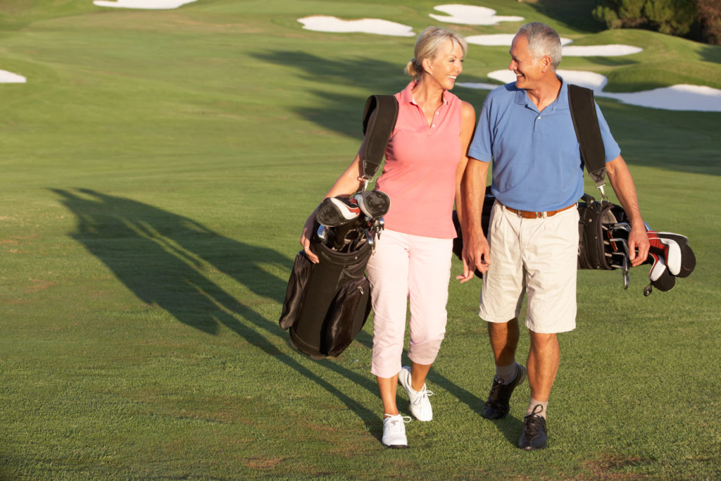 Senior Couple Walking Along Golf Course Carrying Bags And Smiling